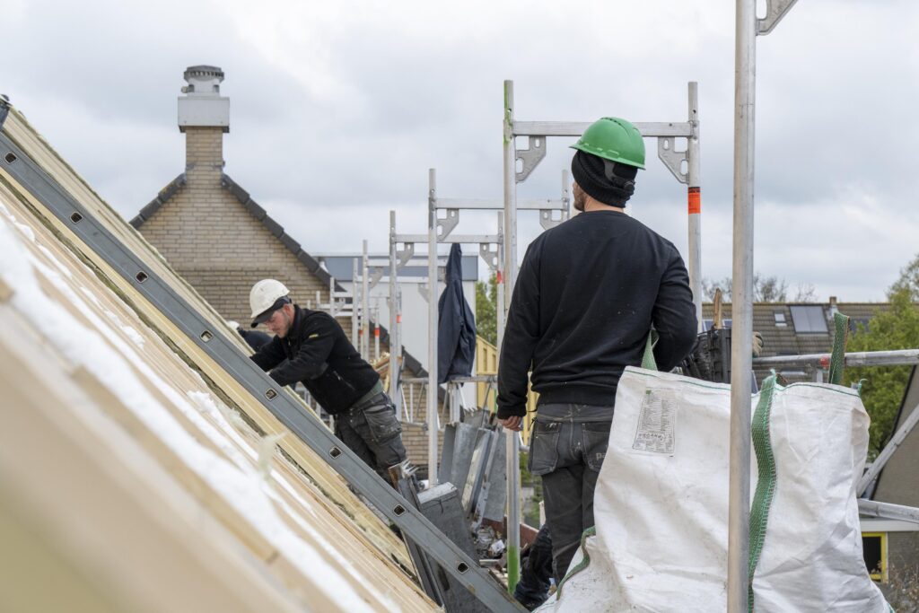 Two construction workers wearing work clothes are installing red roofing on a building.