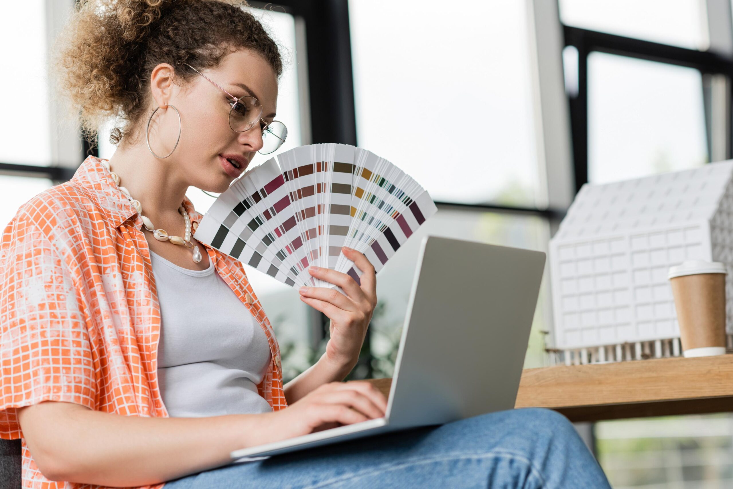 curly architectural designer in glasses holding color palette near laptop in office