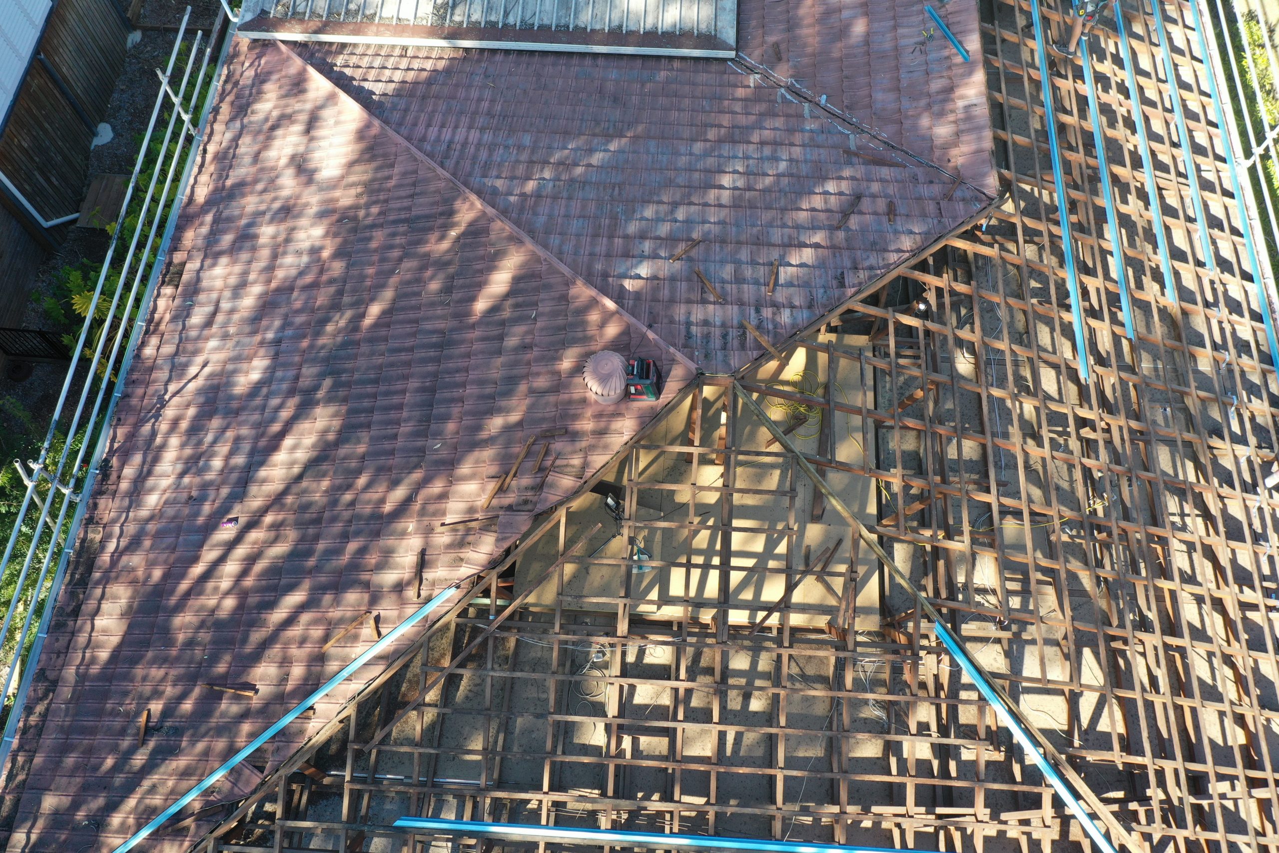 A tile roof being removed, seen from above, with exposed timber battens on one half and intact tiles on the other.